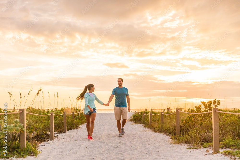 Happy young couple in love walking on romantic beach stroll at sunset. Lovers holding hands on Florida vacation holidays. People walking on summer evening lifestyle.
