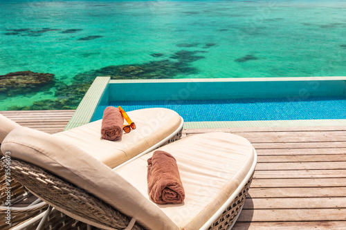 Fototapeta Naklejka Na Ścianę i Meble -  Luxury hotel infinity swimming pool with two sun loungers on deck with towels for couple's honeymoon relaxing overwater bungalow suite. Ocean reefs blue background.