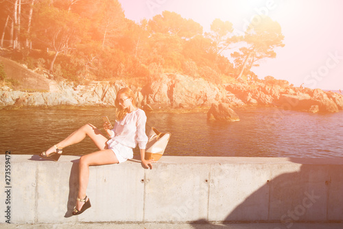 Beautiful caucasian blonde woman enjoy the beautiful seascape of l'esterel region posing glamourously on a rocks and using her smartphone,flare added. photo
