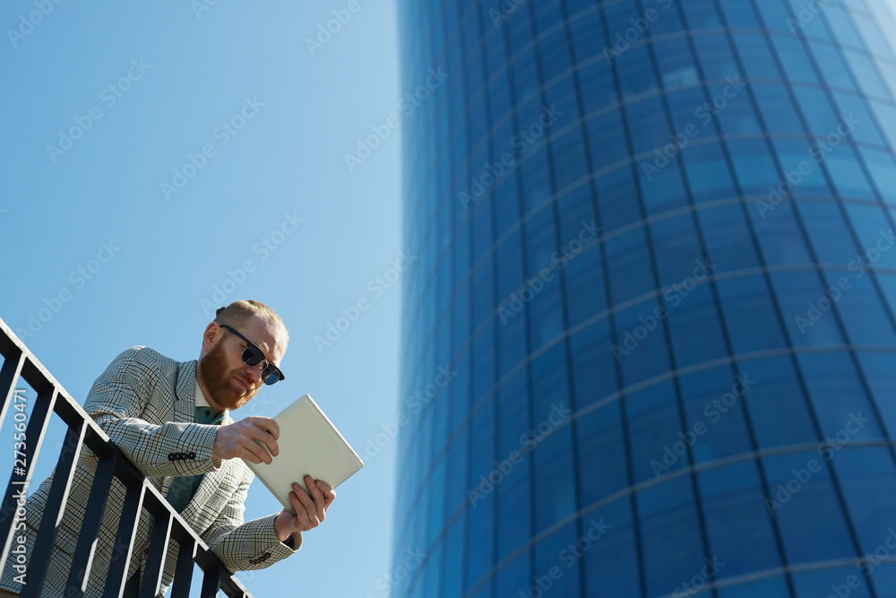 Below view of serious city guy in sunglasses leaning on railing in financial district and working with digital tablet Below view of serious city guy in sunglasses leaning on railing in financial district and working with digital tablet