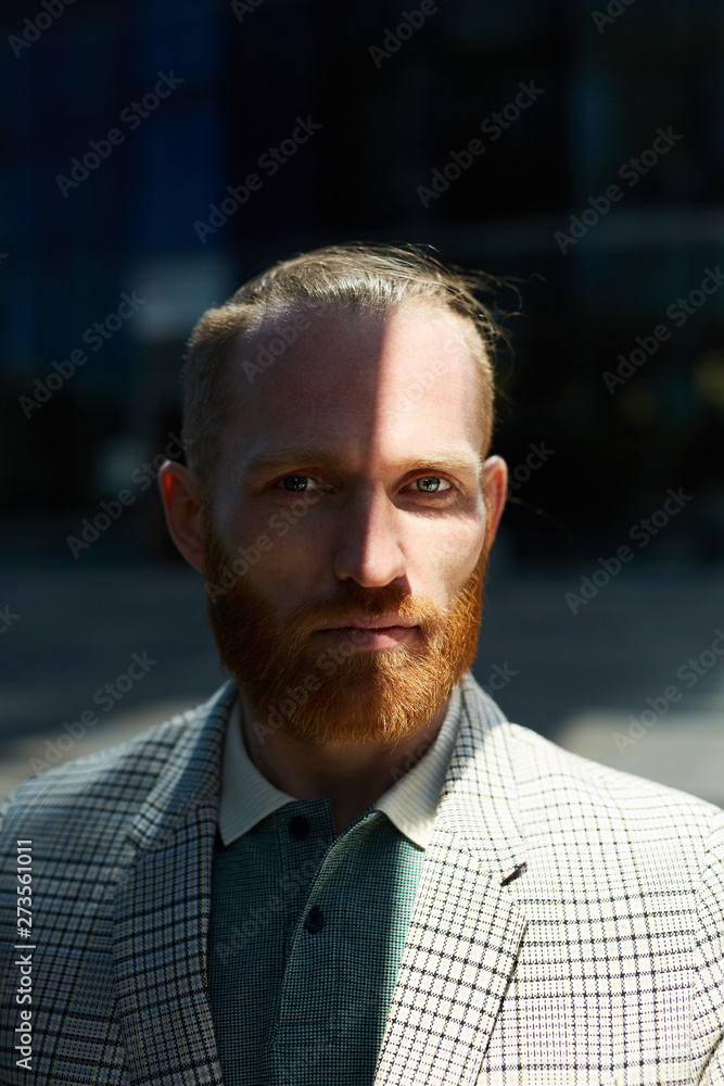 Portrait of serious confident businessman with red mustache and beard wearing fashionable checkered jacket standing in shade outdoors