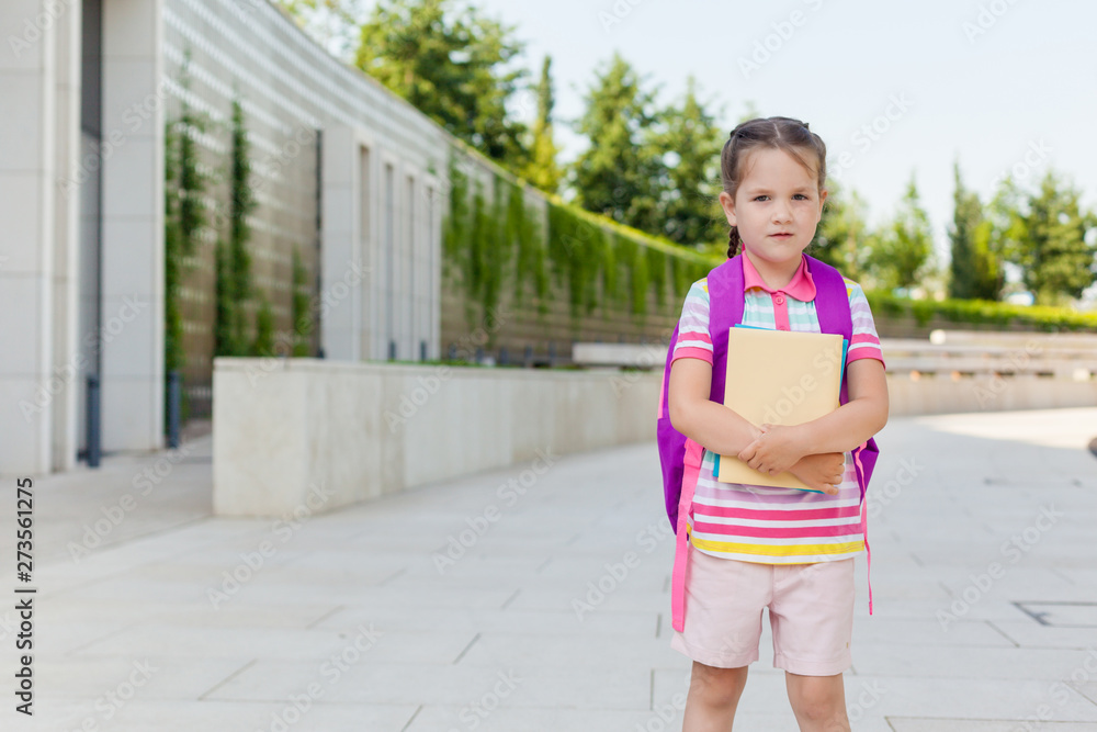 First day of school. Pupil of primary school with book in hand. . Girl with a backpack near the building outdoor. Beginning of classes. The first day of autumn. concept back to school. First day of school. Pupil of primary school with book in hand. . Girl with a backpack near the building outdoor. Beginning of classes. The first day of autumn. concept back to school.