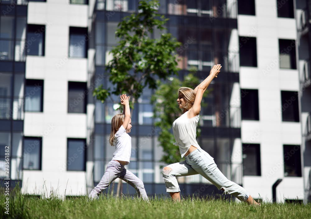Mother and daughter working out practicing yoga outside on a grass together urban city  park 