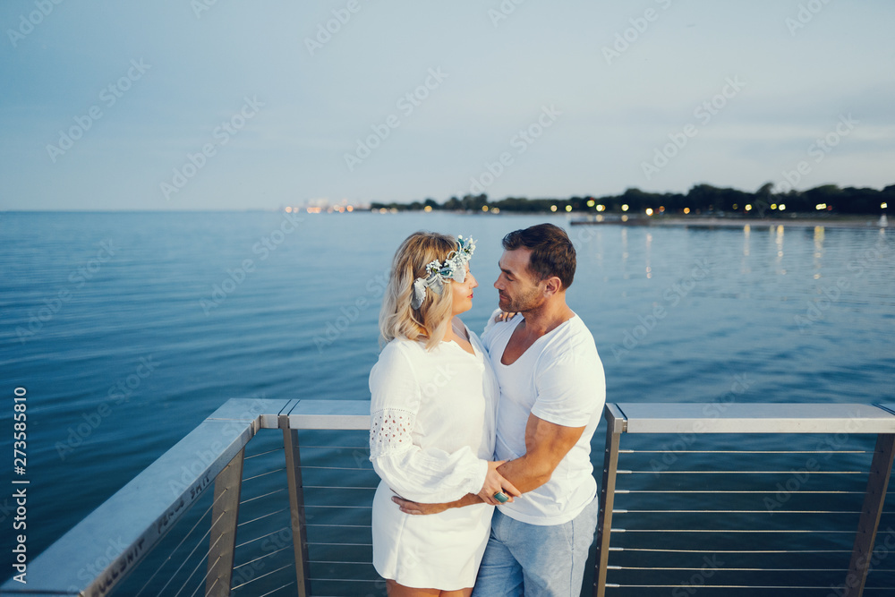 elegant and stylish woman with light curly hair dressed in a white blouse standing near water along with her elegant man in a white t-shirt and in a  blue shorts