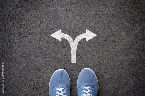 Pair of feet standing on tarmac road with arrow print pointing in two different directions for the concept of making decision at crossroad.