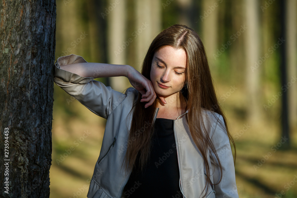 Portrait of a young pretty caucasian teen girl with long hair in casual clothes in the forest on a summer evening sunset.