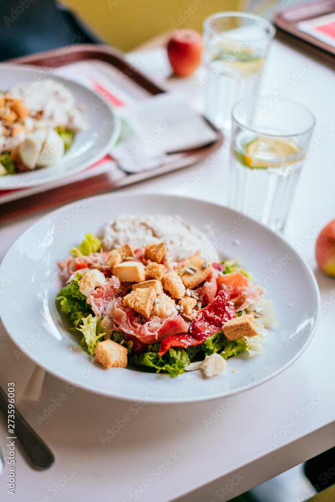 Plate of salad with tomatoes, jamon and baked vegetables on the white table. Glass of iced water with mint and lemon. Plate of salad with tomatoes, jamon and baked vegetables on the white table. Glass of iced water with mint and lemon.