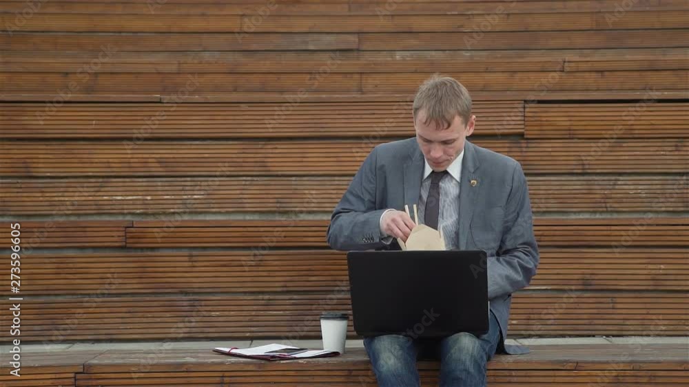 Businessman Using Laptop Outdoors while Having Lunch. He is Eating Chinese Noodles while Working Outside Office. Lifestyle and Business Concept Businessman Using Laptop Outdoors while Having Lunch. He is Eating Chinese Noodles while Working Outside Office. Lifestyle and Business Concept