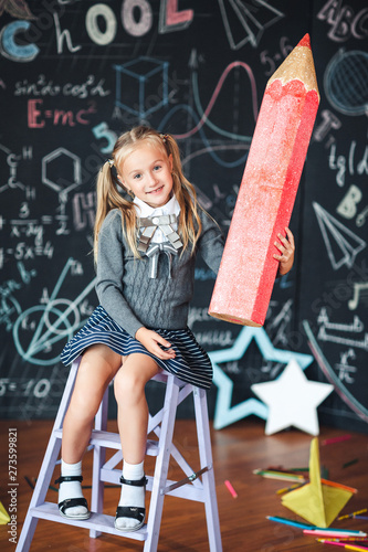 Back to school! A little happy blonde girl in school uniform sits with very large red pencil in her hands at school against chalkboard with school formulas . School Girl responds to the lesson.