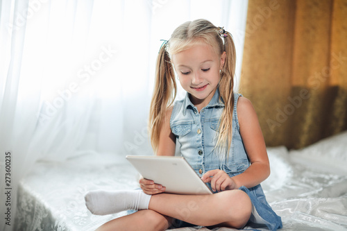 Foto Cute little child girl blond in denim sundress lies in bed uses digital tablet