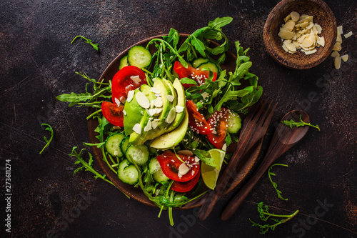 Green salad with avocado, tomatoes, cucumbers and nuts in wooden plate on dark background.