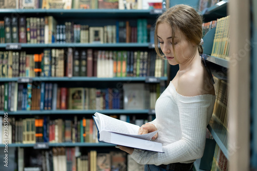 A beautiful young female student is looking for a textbook on the shelves in a bookstore to prepare for a new school year. Literature for learning.