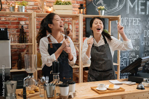 two asian happy girl worker colleagues with aprons enjoy dancing while listening to music in bakery. carefree young women waitress coworkers singing with songs in bar counter in modern coffee store