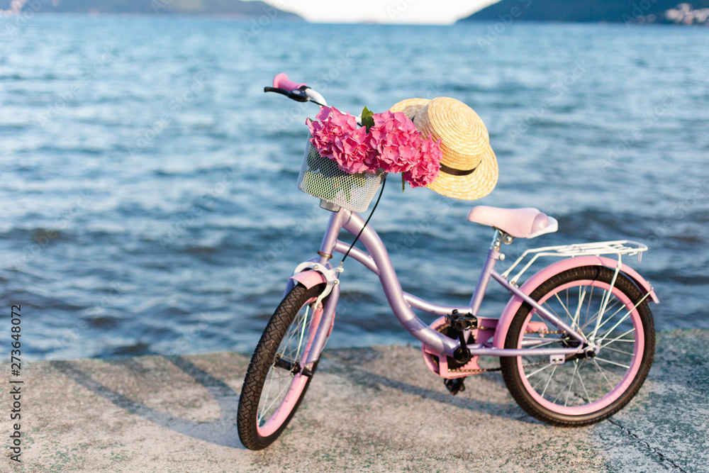 Fototapeta premium Female romantic bicycle with pink hydrangea flowers and straw hat on sea beach. Concept of children riding on coastline, tenderness, childhood, summer.