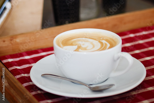 A big cup of freshly brewed cappuccino with latte art heart coffee on wooden table, red plaid tablecloth in cozy craft cafe with shallow depth of field and copy space.