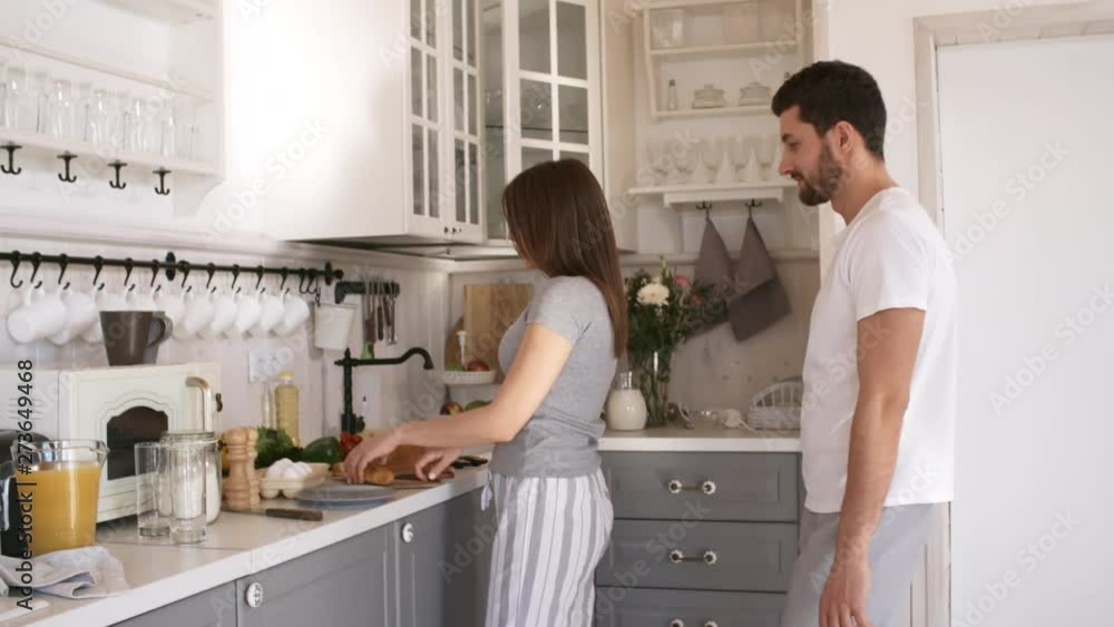 Handheld shot of young woman in sleepwear walking towards kitchen counter and cutting bread in morning as her loving husband hugging her from behind Handheld shot of young woman in sleepwear walking towards kitchen counter and cutting bread in morning as her loving husband hugging her from behind