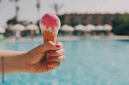 Female hand with a beautiful manicure holds pink ice cream on a hot day on background of a blue pool. The concept of leisure, summer vacation. Copy space. Selective focus.