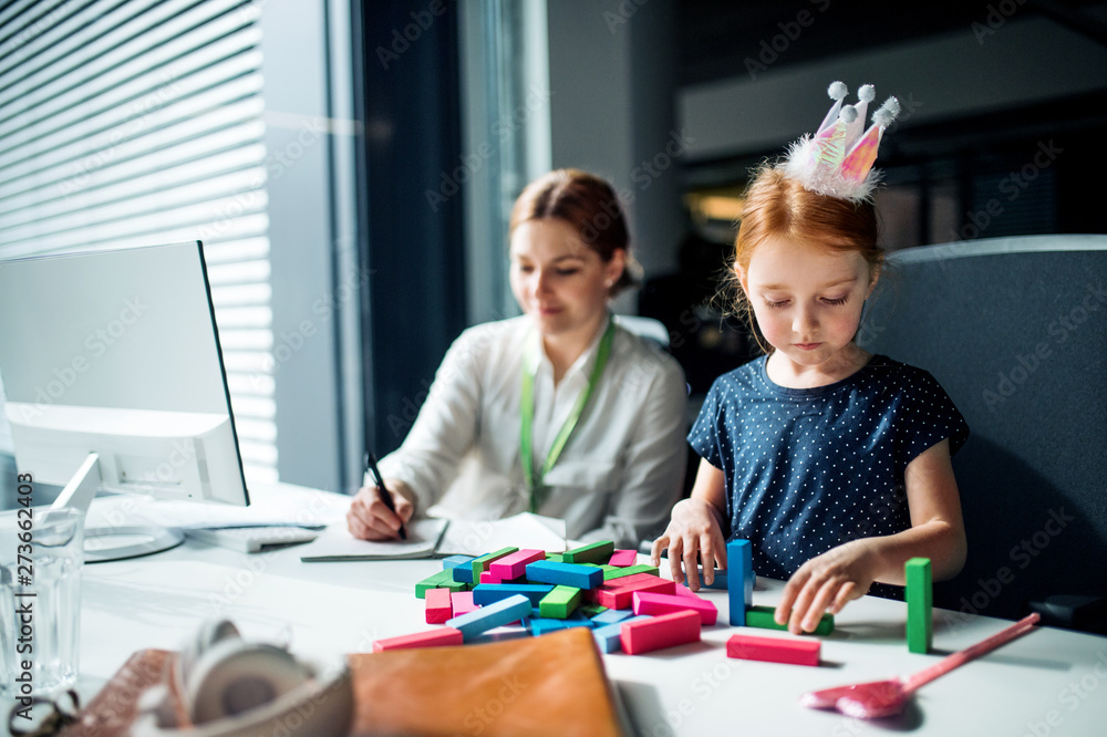 A businesswoman with small daughter sitting in an office, working. A businesswoman with small daughter sitting in an office, working.
