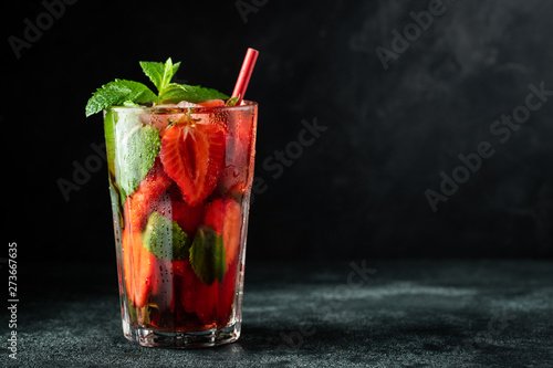 Fresh lemonade with ice, mint and strawberry on top in glass on black table background, copy space. Cold summer drink. Sparkling glasses with berry cocktail
