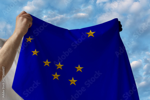 beautiful national flag of alaska in male hands against the blue sky with clouds