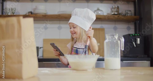 Small girl in cook cap mixes dough in bowl and watches video on smartphone photo