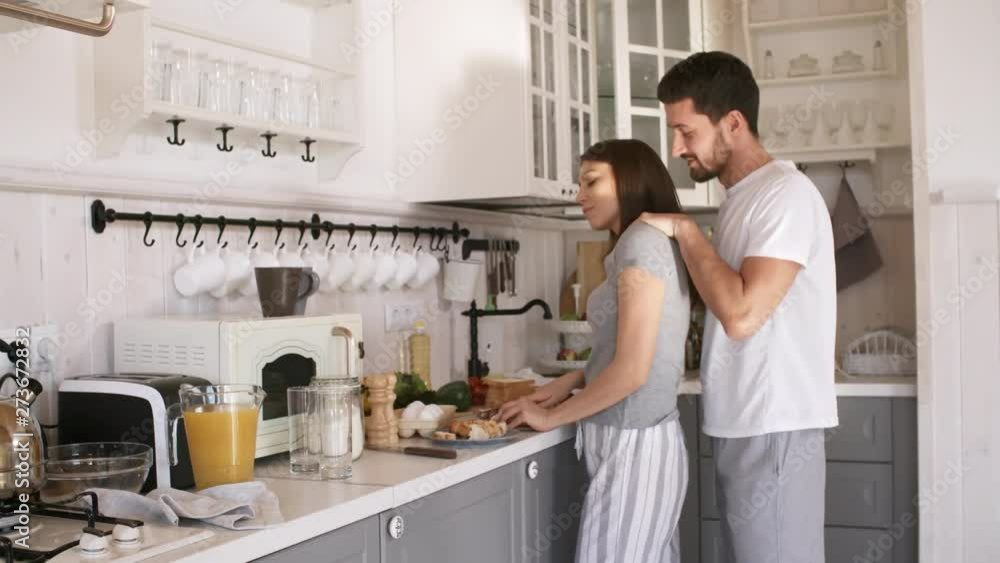 Handheld shot of young woman in sleepwear cutting bread in kitchen and smiling as loving husband caressing her and kissing her on cheek Handheld shot of young woman in sleepwear cutting bread in kitchen and smiling as loving husband caressing her and kissing her on cheek