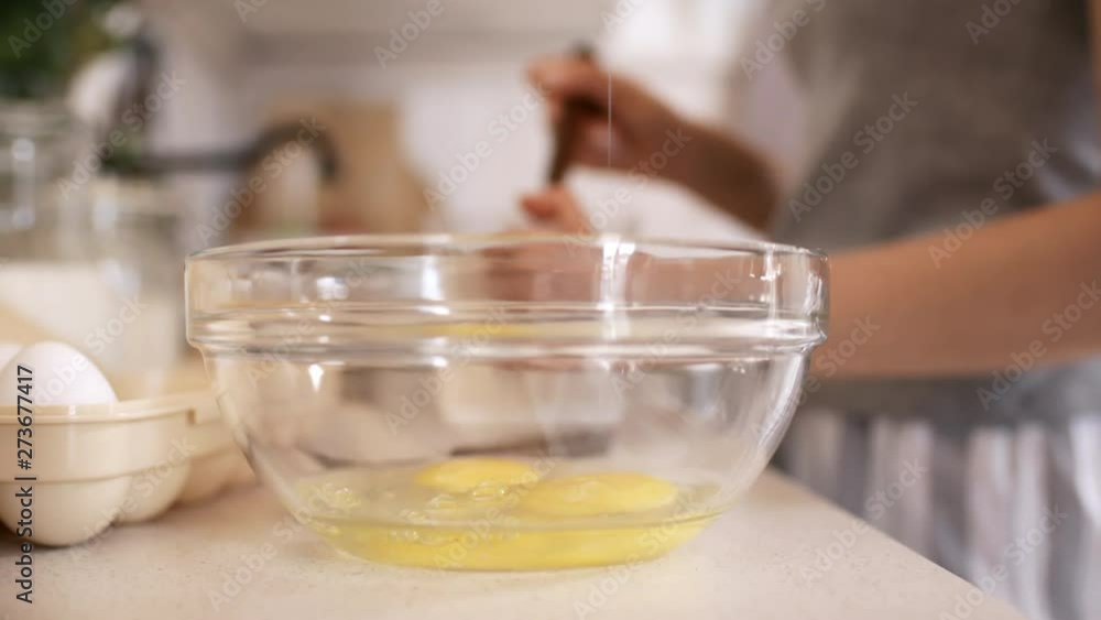 Close up of male hand cracking egg into glass bowl while making breakfast in morning. Unrecognizable woman stirring something in background Close up of male hand cracking egg into glass bowl while making breakfast in morning. Unrecognizable woman stirring something in background