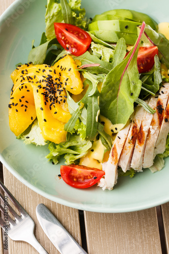 Salad with grilled chicken, mango, lettuce, avocado, tomatoes, arugula, cheese sause on a white plate on wooden background