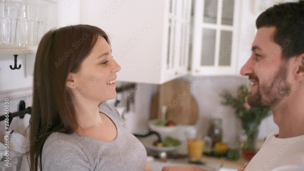 Handheld shot of affectionate man with beard kissing young woman sitting on kitchen counter and smiling Handheld shot of affectionate man with beard kissing young woman sitting on kitchen counter and smiling
