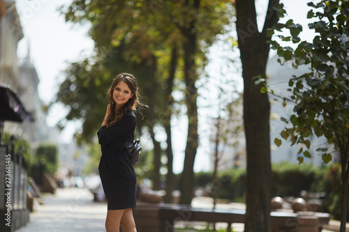 Beautiful and fashionable model girl with a shiny smile in a stylish black short dress walking by the summer city street in sunny day © innarevyako
