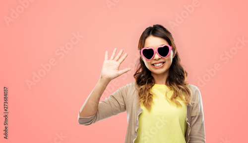 summer, valentine's day and eyewear concept - happy asian young woman in pink heart-shaped sunglasses waving hand over living coral background