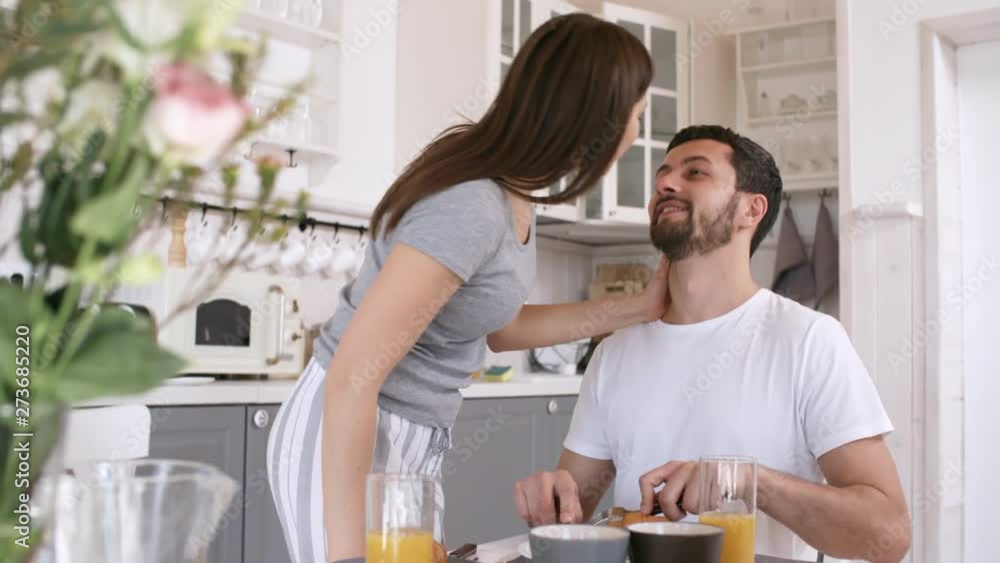 Handheld shot of happy young woman in sleepwear kissing her boyfriend and sitting down at kitchen table to eat breakfast in morning Handheld shot of happy young woman in sleepwear kissing her boyfriend and sitting down at kitchen table to eat breakfast in morning