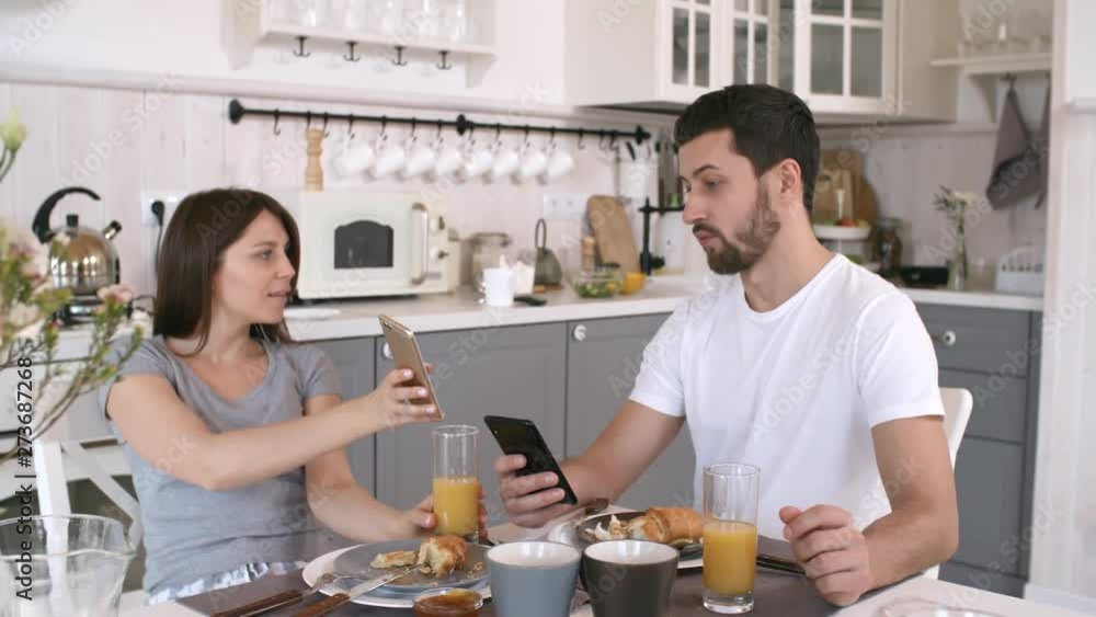 Handheld shot of young woman and bearded man sitting at kitchen table and showing each other funny pictures on mobile phones while having breakfast together Handheld shot of young woman and bearded man sitting at kitchen table and showing each other funny pictures on mobile phones while having breakfast together
