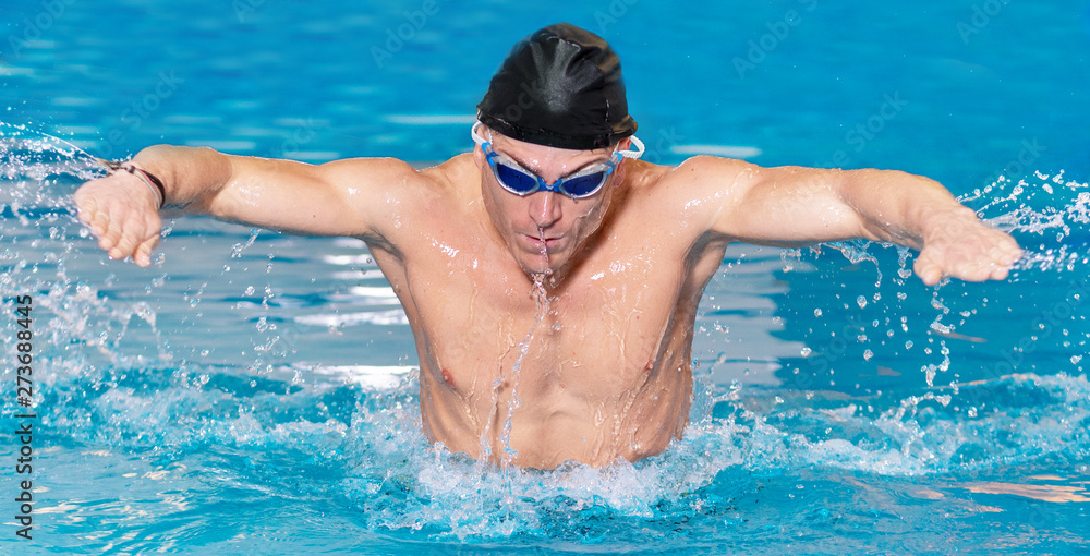Panorama of Muscular swimmer young man in black cap in swimming pool, performing butterfly stroke .