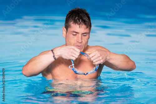 Handsome muscular man standing inside the pool putting on his swimming googles .