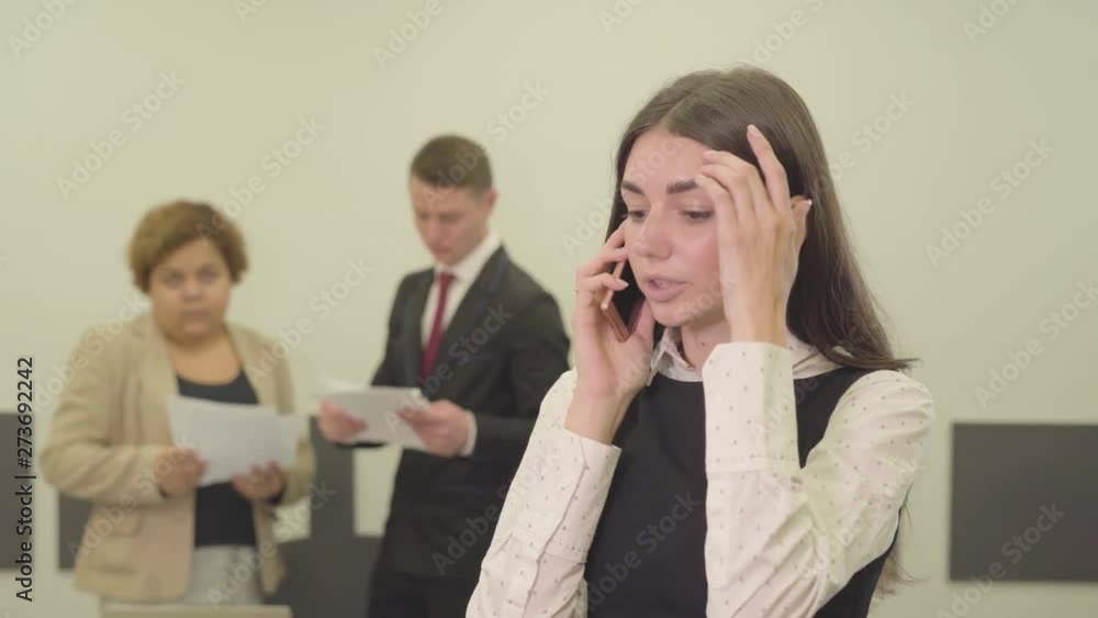 Portrait of adorable attractive young woman in formal wear talking by cellphone in the foreground in the office while her male and female colleague working with papers on the background. Office life Portrait of adorable attractive young woman in formal wear talking by cellphone in the foreground in the office while her male and female colleague working with papers on the background. Office life