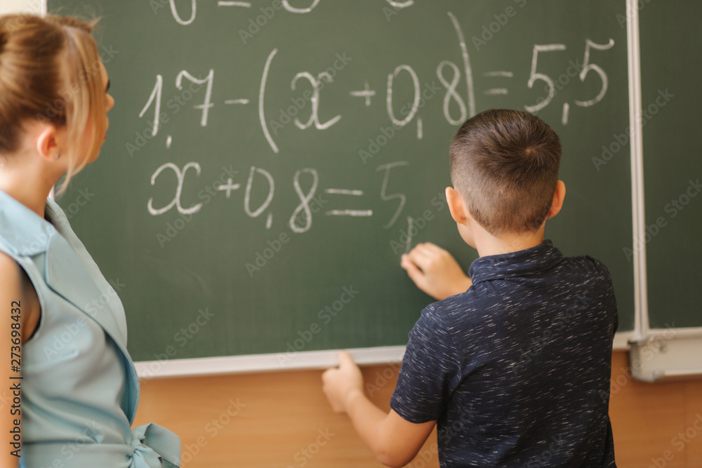 Young female teacher help schoolboy. Boy stand by the blackboard Young female teacher help schoolboy. Boy stand by the blackboard