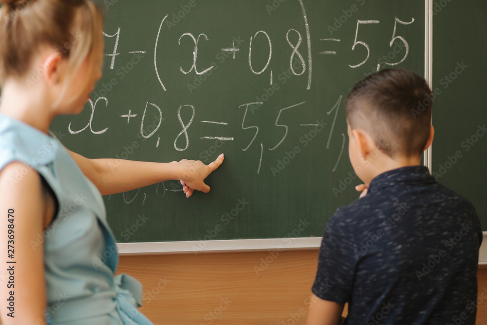 Young female teacher help schoolboy. Boy stand by the blackboard Young female teacher help schoolboy. Boy stand by the blackboard