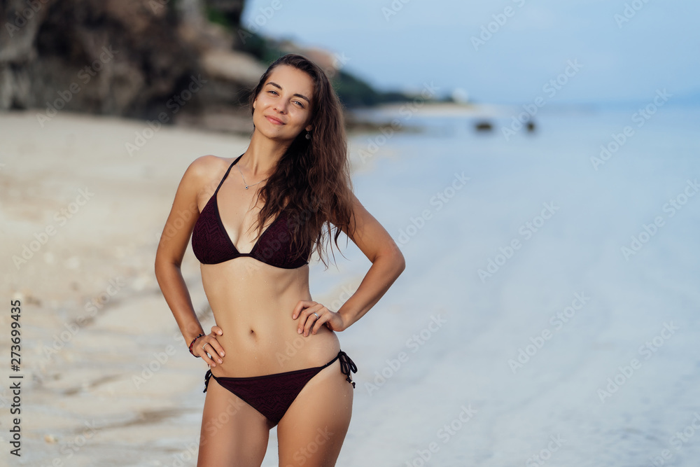 Smiling girl with perfect body in black swimwear posing on white sand beach