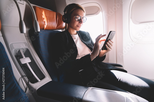 Young caucasian woman sitting near window with smartphone connected to wifi internet during flight on board, female passenger in headphones for noise cancellation search songs in online shop on app