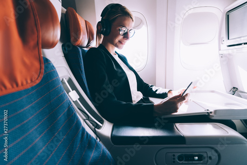 Happy smiling woman passenger in headphones for noise cancellation enjoying intercontinental flight in first class with high speed wifi connection on board using touch pad and browsing internet photo