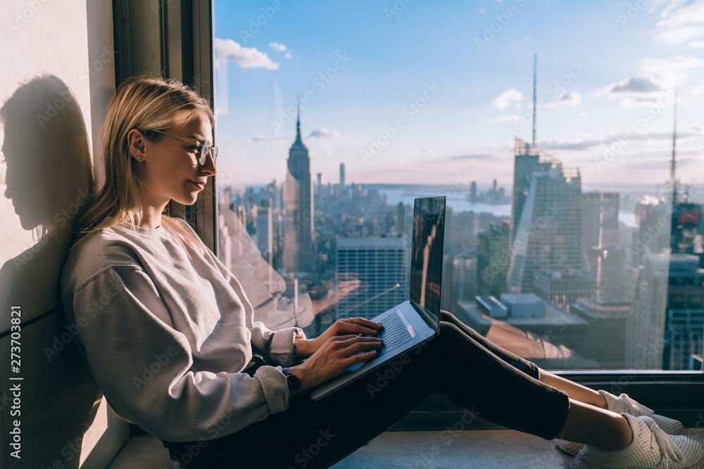 Fototapeta premium Caucasian female digital nomad keyboarding article for travel blog while working on laptop computer in modern coworking office with panoramic views of New York downtown. Woman tourist at window sill