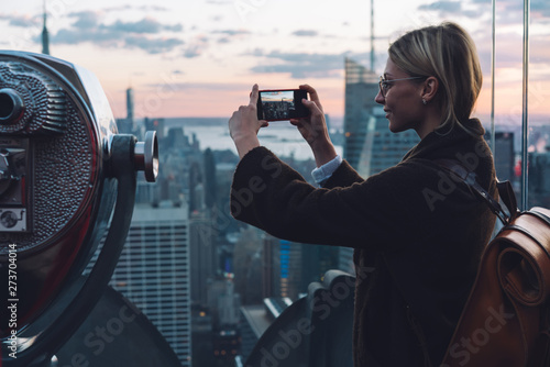 Fototapeta Caucasian young woman traveler making video on cellphone camera while standing on open Observation Deck with scenery view New York cityscape at sunset