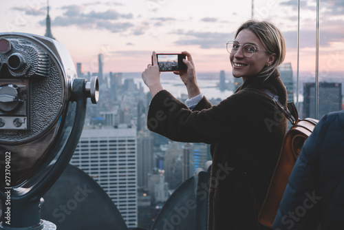 Happy carefree female tourist taking photo on smartphone camera while visiting famous Observation deck with scenery view of New York landmarks. Hipster girl enjoying her ravel to USA during vacations