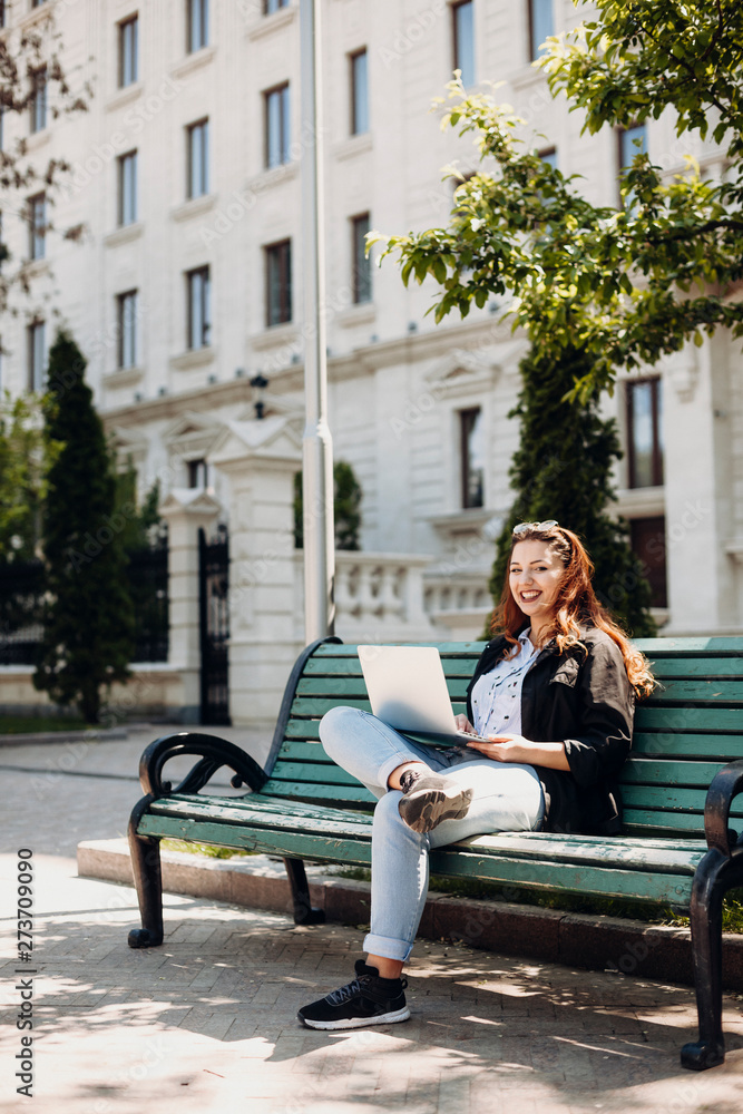 Full length portrait of a plus size woman sitting on a bench against a building with a laptop on her legs looking at camera laughing.