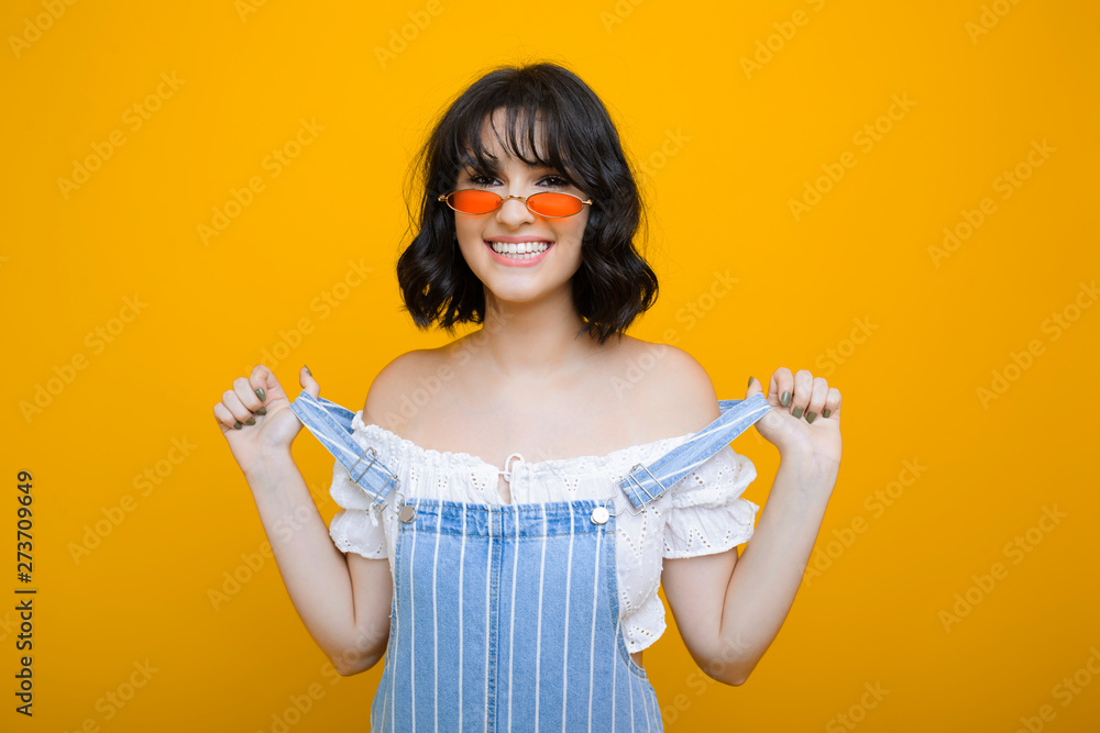 Beautiful young caucasian woman with a short hair looking at camera while laughing while holding her jeans dress in front of a yellow wall. Beautiful young caucasian woman with a short hair looking at camera while laughing while holding her jeans dress in front of a yellow wall.