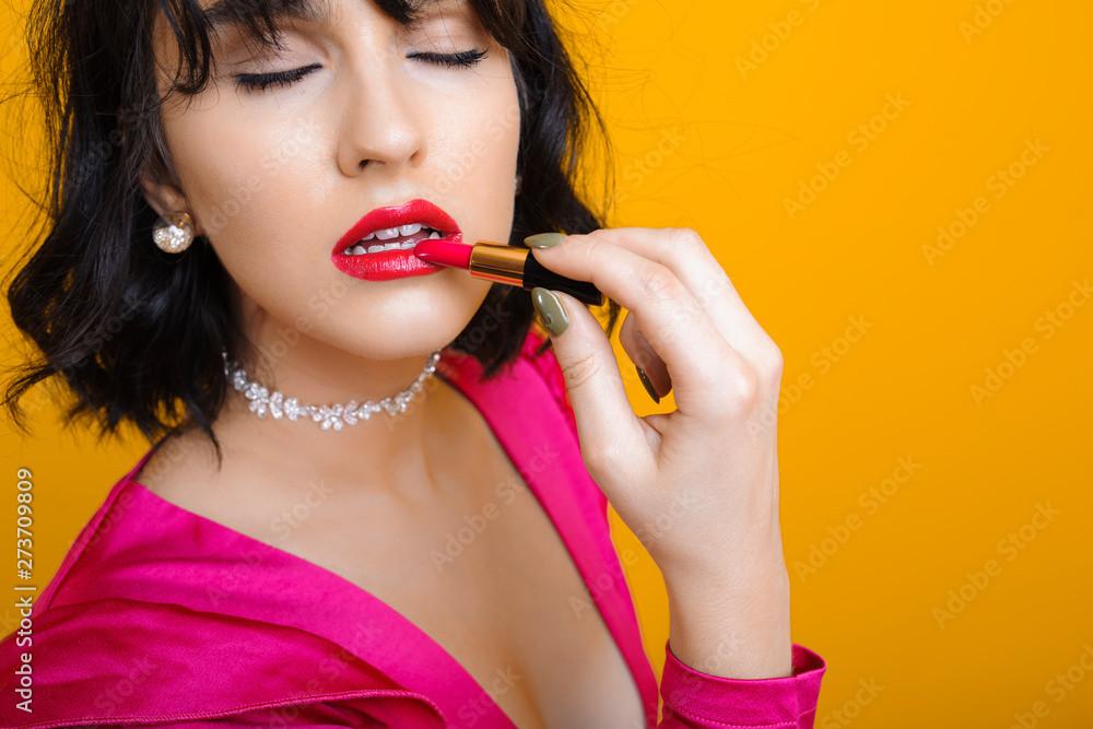 Close up portrait of a beautiful female influencer holding a red lipstick on her lips with closed eyes dressed in a pink dress against a yellow wall. Close up portrait of a beautiful female influencer holding a red lipstick on her lips with closed eyes dressed in a pink dress against a yellow wall.