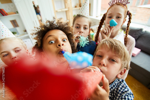 High angle view at multi ethnic group of children blowing party horns at camera and having fun