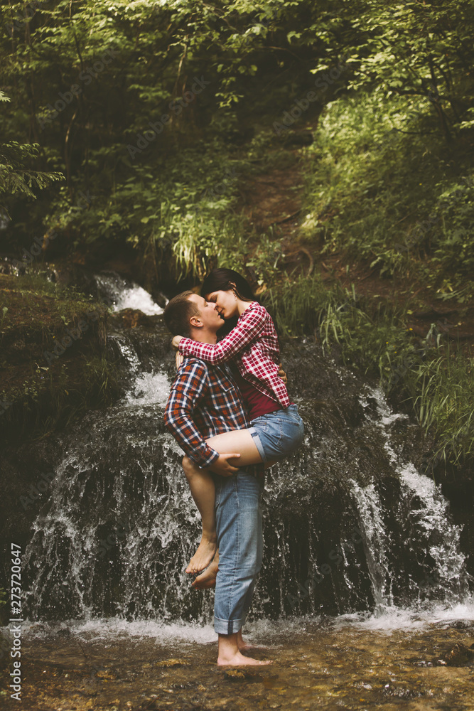 Beautiful young couple kisses against the backdrop of an amazing waterfall. Man and woman are dressed in jeans and plaid shirts. Concept of travel and unusual dates.