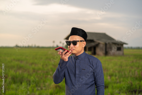 Portrait of handsome young Malay Asian man in Baju Melayu using smartphone, texting, voice mail, talking, making a call with traditional house, paddy field meadow background